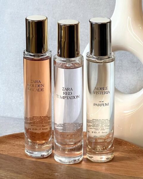 Trzy flakony ikoniczne perfumy z Zary w małym rozmiarze podróżnym (mini), ustawione na drewnianej tacy. Od lewej: Zara Golden Decade, Zara Red Temptation i Zara Noble Wisteria Eau de Parfum. Butelki mają złote nakrętki, a płyny wewnątrz są w odcieniach od brzoskwiniowego (Golden Decade) po przezroczysty/jasny (Red Temptation i Noble Wisteria). W tle znajduje się biała, ceramiczna waza.