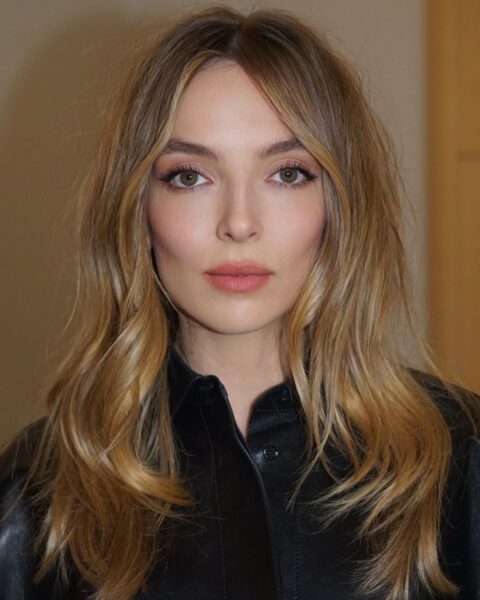 Jodie Comer z idealnym makijażem „smoky eyes” i fryzurą w stylu fal „beach waves”. Gwiazda pozuje na pokazie Givenchy w czarnej skórzanej koszuli. Doskonała inspiracja, jak uzyskać promienny wygląd za pomocą fal i naturalnego makijażu, który podkreśla rysy twarzy.