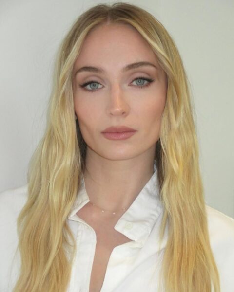 Zbliżenie na aktorkę Sophie Turner w klasycznej, białej koszuli. Ma długie, proste włosy w odcieniu ciepłego blondu z subtelnym, naturalnym balejażem i przedziałkiem na środku. Delikatny makijaż z akcentem na perfekcyjnie wyciągniętą, czarną kreskę na powiece i nude'owe usta podkreślają jej błękitne oczy. Inspiracja do stworzenia minimalistycznego, eleganckiego looku.