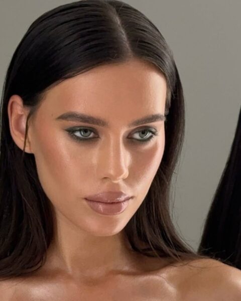 Portret pięknej kobiety o ciemnych włosach i wyrazistym, wieczorowym makijażu. Jej spojrzenie jest intensywne dzięki smoky eye w kolorze głębokiej, przydymionej zieleni lub grafitu, z idealnie roztartą kreską na dolnej powiece. Cera jest perfekcyjnie wykonturowana, z efektem zdrowej opalenizny i naturalnego blasku, a usta muśnięte błyszczykiem w odcieniu nude. Ciemne włosy są gładko zaczesane z przedziałkiem na środku. Zdjęcie ilustruje wyrafinowany look, idealny jako inspiracja do makijażu wieczorowego dla kobiet ceniących klasę i elegancję.