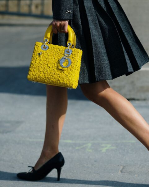 Żółta torebka Dior Lady Dior (lub Lady D-Lite) z fakturowanej skóry z kultowymi metalowymi charmsami D.I.O.R., trzymana w dłoni. Stylizacja obejmuje czarną, plisowaną spódnicę i czarne, spiczaste czółenka na wysokim obcasie.