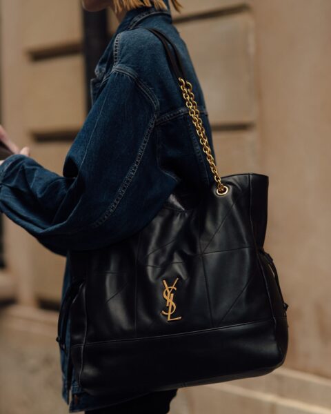 Duża, czarna torebka-worek (shopper) Saint Laurent Icare Maxi Shopping Bag z pikowanej skóry z charakterystycznym, złotym logo YSL. Torebka zawieszona na ramieniu osoby ubranej w oversize'ową kurtkę dżinsową w ciemnym kolorze.