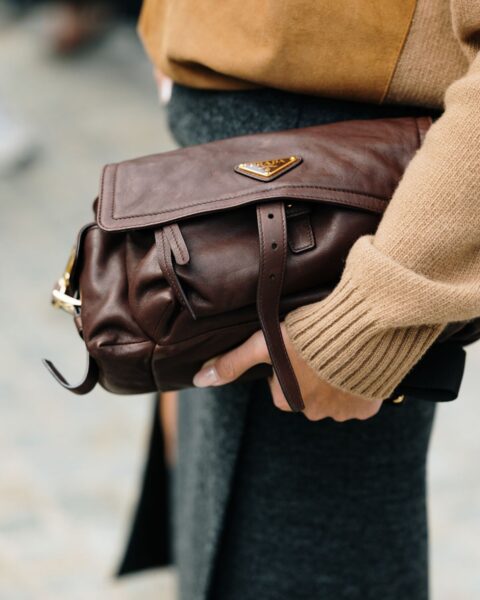 Brązowa, skórzana torebka na ramię Prada (prawdopodobnie model Re-Edition lub podobny fason vintage) z klapą i charakterystycznym trójkątnym logo, trzymana przy boku. W tle widoczny karmelowy, wełniany sweter i grafitowa spódnica midi z rozporkiem. Zdjęcie ilustruje temat: jak inwestować w torebki.