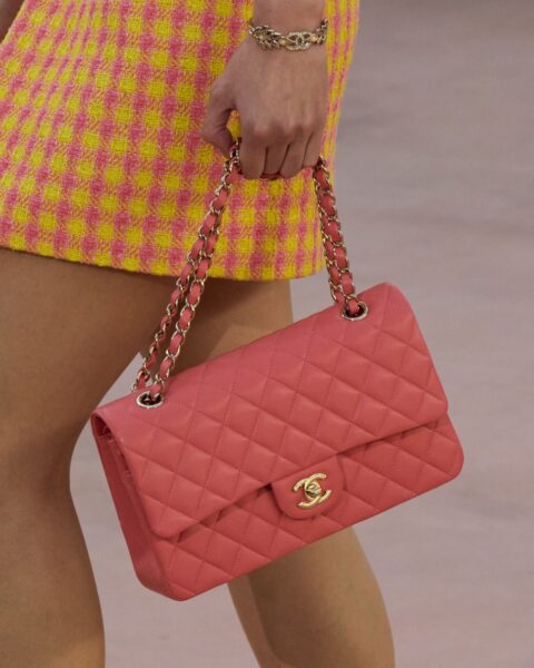Mała pikowana torebka Chanel 2.55 (lub Classic Flap Bag) w intensywnym kolorze koralowej czerwieni/różu, ze złotym łańcuchem i kultowym zapięciem CC. Torebkę trzyma modelka ubrana w mini spódnicę z tweedu w wyrazistą, żółto-różową kratę. Na nadgarstku widoczna złota bransoletka-łańcuch.