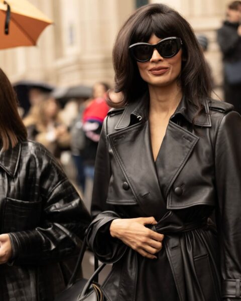 Zdjęcie street style z Tygodnia Mody w Paryżu: Kobieta o ciemnych włosach w stylowym, czarnym trenczu ze skóry (lub imitacji skóry) z paskiem, dużych okularach przeciwsłonecznych typu cat-eye oraz z małą, czarną torebką. Elegancki i wyrazisty look idealny na deszczowy dzień, pokazujący, jak klasyczny fason płaszcza może zyskać rockowy charakter dzięki skórzanej fakturze. Moda jesień-zima 2025/2026.