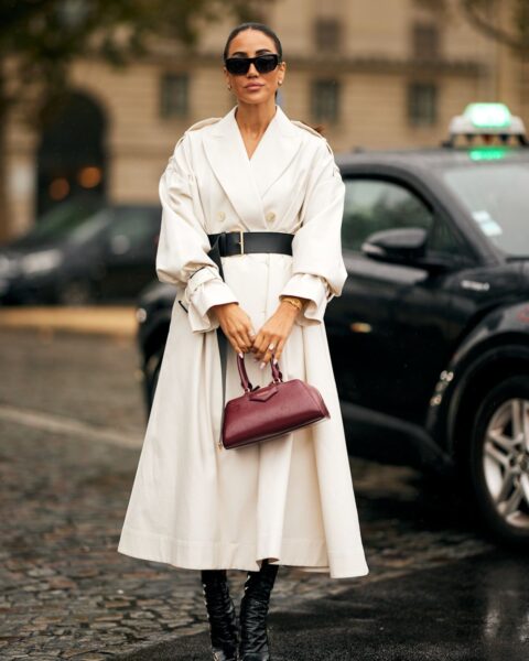 Zdjęcie street style z Paryża: Kobieta w eleganckim, długim trenczu w kolorze écru lub złamanej bieli, podkreślającym talię szerokim, czarnym paskiem. Stylizację uzupełniają czarne, dopasowane botki na obcasie i wyrazista torebka w odcieniu głębokiego bordo lub wiśni. Akcesoria to czarne okulary przeciwsłoneczne i delikatne złote bransoletki. W tle widać ulicę i taksówkę. To inspirujący look na jesień, który łączy klasyczną formę trencza z nowoczesną, wyrafinowaną paletą barw i akcentami.