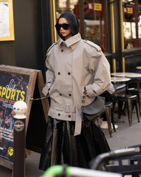 Zdjęcie street style z Tygodnia Mody w Paryżu: Kobieta w nowoczesnym, beżowym, krótkim trenczu o oversize'owym kroju, z obszernymi rękawami. Pod spodem czarna spódnica midi z błyszczącego lub plisowanego materiału oraz czarny kaptur lub kominiarka. Stylizację uzupełniają duże czarne okulary przeciwsłoneczne i czarna torebka-półksiężyc na łańcuchu. Inspirujący przykład łączenia sportowych i eleganckich elementów oraz nowoczesnej interpretacji klasycznego trencza.
