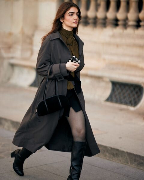 Zdjęcie street style z Tygodnia Mody w Paryżu: Kobieta w długim trenczu w odcieniu ciemnego grafitu lub taupe. Płaszcz narzucony jest na stylizację z oliwkową bluzką lub apaszką z wiązaniem pod szyją i krótką, czarną spódniczką. Całość uzupełniają kryjące czarne rajstopy oraz wysokie, czarne kozaki na masywnym obcasie. W ręku mała, czarna torebka typu baguette. Elegancki i klasyczny look z nowoczesnym akcentem kolorystycznym, idealny na chłodniejsze dni, inspirujący do tworzenia zestawów z klasą.