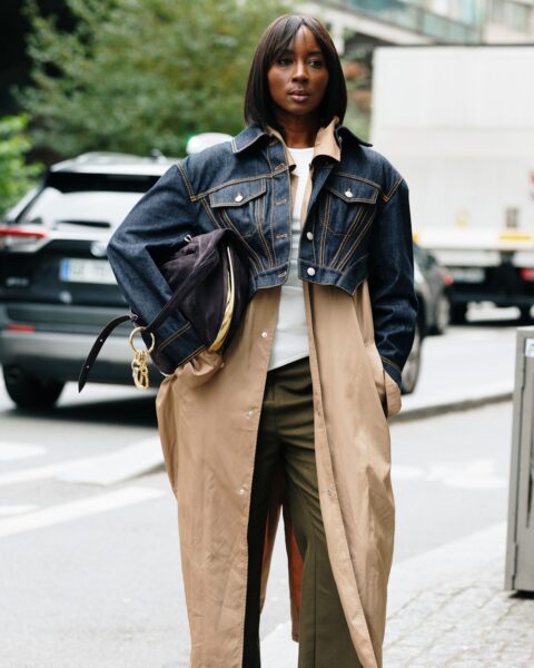 Zdjęcie street style z Tygodnia Mody w Paryżu: Kobieta w warstwowej stylizacji. Na sobie ma długi, luźny, beżowy płaszcz typu trencz lub prochowiec, na który narzucona jest krótka kurtka jeansowa z ciemnego denimu z wywiniętym kołnierzem. Pod spodem jasna bluzka i szerokie, oliwkowe spodnie. W ręce trzyma brązową torebkę-worek ze złotymi detalami. Uwagę zwracają oryginalne, żółte buty z futrzanymi elementami. Nowoczesne, eklektyczne podejście do łączenia klasycznych elementów z awangardowymi detalami.