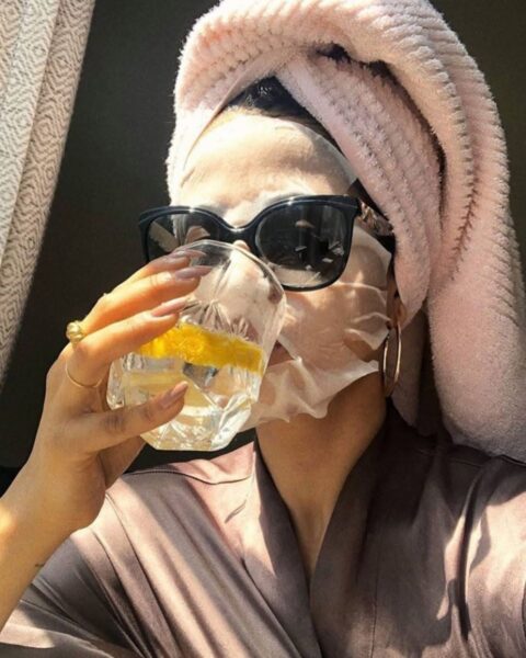 Zbliżenie na kobietę w luksusowym, satynowym szlafroku i różowym turbanie na głowie, która ma na twarzy nałożoną maseczkę w płachcie. Na nosie ma duże, czarne okulary przeciwsłoneczne, a w ręce trzyma szklankę z napojem z plastrem cytryny, pijąc lub zbliżając ją do ust. Zdjęcie w ciepłym, słonecznym świetle wpadającym przez okno.