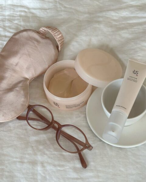 Na białej, pogniecionej tkaninie (prawdopodobnie pościeli) leżą akcesoria i kosmetyki do pielęgnacji: jasnoróżowa, satynowa opaska na oczy do spania, brązowe okulary korekcyjne z przezroczystymi oprawkami, otwarte beżowe pudełko z płatkami pod oczy (Revive Under Eye Patch) i tubka kremu (Ginseng Cleansing Oil) firmy Beauty of Joseon. Obok kremu znajduje się mała biała filiżanka ze spodkiem. To ujęcie typu flat lay przedstawiające produkty do koreańskiej pielęgnacji i relaksu.