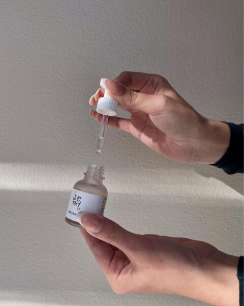 Dwie dłonie na neutralnym, jasnym tle trzymają małą, szklaną buteleczkę z pipetą (droperem) z korejskiem serum do twarzy marki Beauty of Joseon. Jedna dłoń trzyma butelkę, druga unosi pipetę, z której spływa kropla kosmetyku. Dłonie mają zadbane, jasne paznokcie. Cień rzucany na ścianę dodaje głębi.