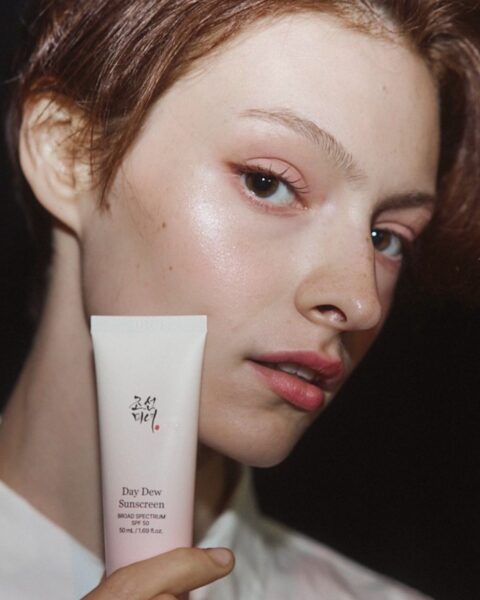 Zbliżenie na twarz kobiety o krótkich, brązowych włosach, z naturalnym, promiennym makijażem, z widocznymi piegami i lśniącą skórą. Kobieta trzyma w dłoni tubkę różowego kremu przeciwsłonecznego Day Dew Sunscreen SPF 50 marki Beauty of Joseon. Ma na sobie białą koszulę. Tło jest ciemne.