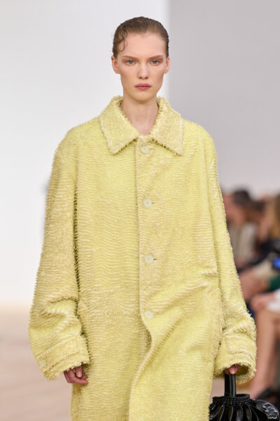 Trendy wiosna 2026: Modelka w pastelowym, żółtym płaszczu z fakturowanej tkaniny o miękkim, welurowym wykończeniu. Oversize’owy krój i subtelna gra światła na materiale podkreślają sensualny minimalizm i nowoczesne krawiectwo domu Loewe.