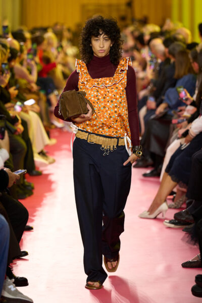 Model w szerokich granatowych spodniach i bordowym golfie prezentuje pomarańczowy fartuszek w kwiaty z białą lamówką i falbankami. Trzyma brązową, pudełkową torbę. Pas z dużą klamrą i przypiętymi elementami nadaje całości użytkowego charakteru, kontrastującego z nawiązaniami do codziennego ubioru wiejskiego.