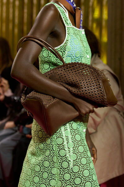 Modelka w miętowej sukience z koronki o kwiatowym wzorze trzyma dużą, brązową, plecioną torbę ze skóry. Naturalna faktura i ciepły kolor akcentują powrót do rzemieślniczych detali i retrofuturystycznego minimalizmu.
