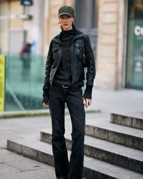 Street style: kobieta w monochromatycznej, czarnej stylizacji w duchu militarnej elegancji. Ma na sobie czarną, skórzaną kurtkę pilotkę, ciemny golf, czarne dżinsy typu bootcut oraz zieloną, kaszkietową czapkę, stojąc na schodach.