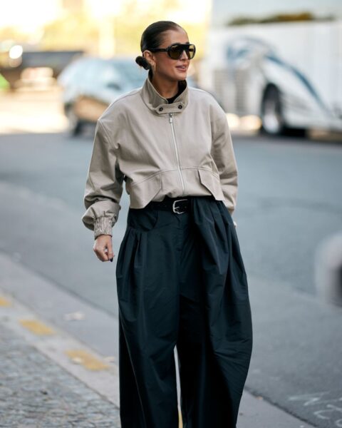 Street style: kobieta w minimalistycznej stylizacji smart casual, łączącej beżową, krótką kurtkę typu bomber z obszernymi, czarnymi spodniami z wysokim stanem (oversize). Ma gładko zaczesane włosy w kok i duże, prostokątne okulary przeciwsłoneczne.