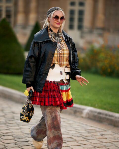 Street style: kobieta w odważnym, eklektycznym zestawieniu boho-punk. Ma na sobie czarną, błyszczącą kurtkę, koszulę w kratę, czerwoną spódnicę mini w szkocką kratę, szeroki biały pas-gorset oraz spodnie z efektem tie-dye. Całość uzupełnia opaska na włosy, naszyjniki typu chunky i okrągłe okulary przeciwsłoneczne.