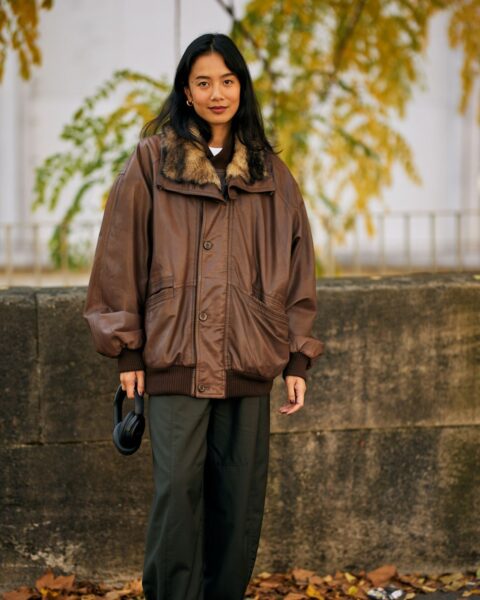 Street style: kobieta w jesiennej stylizacji, nosząca brązową, skórzaną kurtkę typu bomber w rozmiarze oversize, z dużym kołnierzem z futerka, oraz ciemnozielone, szerokie spodnie. Całość uzupełniają czarne botki i złote kolczyki. W tle widać jesienne liście.