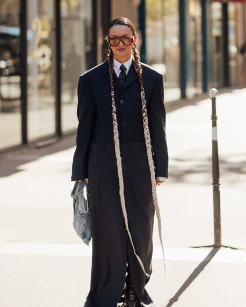 Street style: kobieta w mocnej, modowej stylizacji, nosząca długi, czarny, prążkowany garnitur w stylu oversize, z krawatem i białą koszulą. Całość uzupełniają długie warkocze, okulary w pomarańczowej oprawce i torebka w kolorze spranego dżinsu.