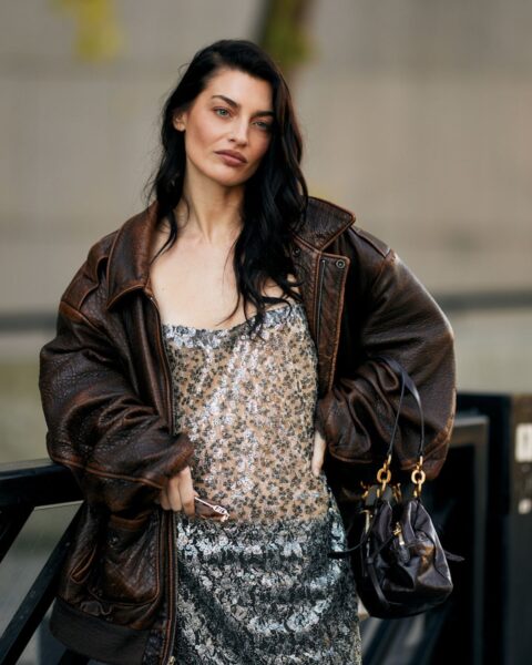 Street style: kobieta w wieczorowej sukience na ramiączkach, pokrytej srebrnymi cekinami, złagodzonej przez dużą, brązową, skórzaną kurtkę pilotkę w stylu vintage (oversize). Całość uzupełnia mała, ciemnobrązowa torebka na łańcuszku.