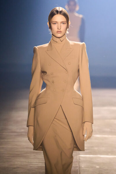 Modelka w camelowym komplecie o rzeźbiarskiej linii ramion, dopasowanym żakiecie i spódnicy o perfekcyjnej konstrukcji. Sylwetka utrzymana w duchu power dressing — dramatyczne światło i ciepły beż tworzą aurę mocy i elegancji.