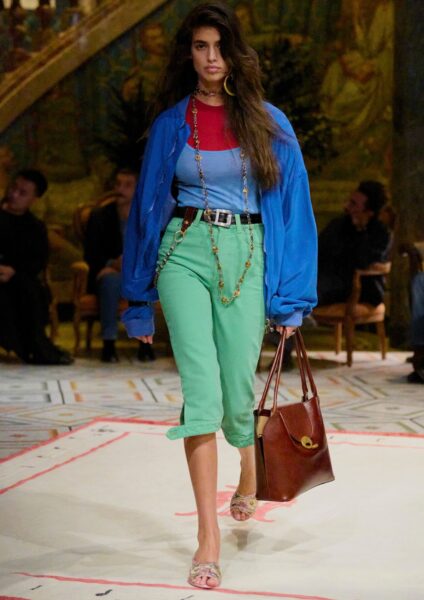 Modelka na wybiegu Versace w looku z kolekcji Pre-Fall 2026. Ma na sobie wielobarwną, warstwową stylizację, na którą składają się: oversizowa, niebieska kurtka lub koszula, niebieski top na ramiączkach i czerwony top z długim rękawem pod spodem, oraz miętowe spodnie rybaczki z podwiniętą nogawką. Całość jest przepasana brązowym paskiem. Stylizację dopełniają: masywne naszyjniki, duże, żółte kolczyki koła, duża brązowa skórzana torebka i klapki na szpilce z ozdobnymi kamieniami.