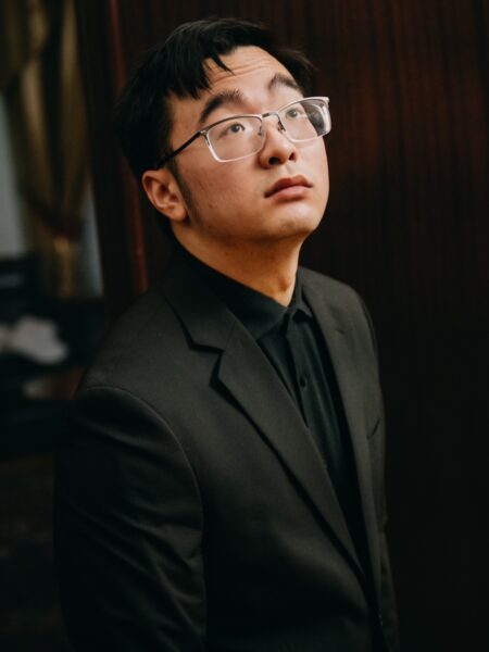 Pianista w czarnym garniturze i okularach patrzy w górę w skupieniu. Zdjęcie wykonane w półmroku ukazuje jego refleksję i napięcie przed występem w Konkursie Chopinowskim.