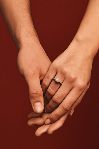 Ponadczasowy blask: Zbliżenie na splecione dłonie kobiety i mężczyzny na tle głębokiej czerwieni – kobieta nosi delikatny pierścionek zaręczynowy z pojedynczym diamentem osadzonym w złotej obrączce, symbolizujący miłość i obietnicę.