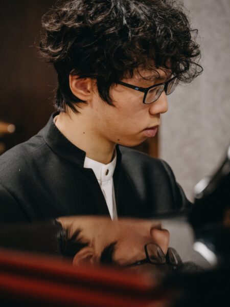 Pianista z kręconymi włosami w okularach gra przy fortepianie w sali prób. Na jego twarzy widać pełne skupienie, a odbicie w lakierowanej powierzchni instrumentu tworzy efekt artystycznego duplikatu.