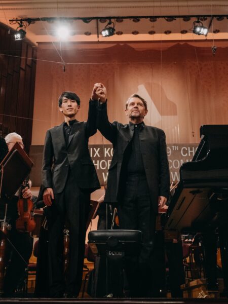 Eric Lu i dyrygent trzymają się za ręce na scenie Filharmonii Narodowej po finałowym występie XIX Konkursu Chopinowskiego. W tle orkiestra i rozświetlony napis z nazwą wydarzenia, a w powietrzu – napięcie, ulga i cisza po ostatnim akordzie koncertu f-moll.