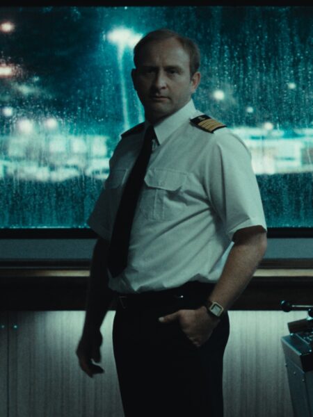 Mężczyzna w białej koszuli z epoletami, czarnym krawacie i zegarku z metalową bransoletą stoi w sterówce statku na tle zaparowanego okna z nocnym pejzażem portu. Ujęcie utrzymane w filmowym, melancholijnym tonie.
