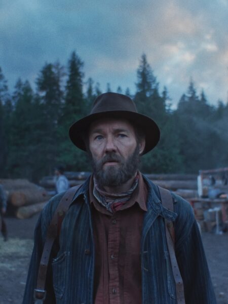 Portret mężczyzny w kapeluszu, koszuli i roboczym fartuchu, stojącego na tle lasu i stosów drewna. Surowe światło poranka i zamglone niebo tworzą melancholijny klimat westernu o samotności i ciężkiej pracy.