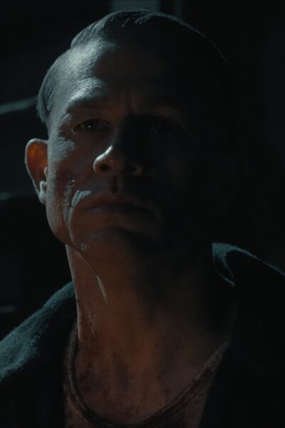 "Potwór: Historia Eda Geina": Mroczny kadr przedstawiający mężczyznę w półcieniu. Jego twarz częściowo oświetlona, pokryta śladami krwi. Patrzy przed siebie z beznamiętnym, zimnym wyrazem twarzy, co potęguje atmosferę niepokoju.