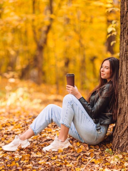 Kolorowe stylizacje: Kobieta siedząca pod drzewem w piękną i złocistą jesień. Ma na sobie skórzaną ramoneskę, jeansy w kolorze baby blue oraz trampki.