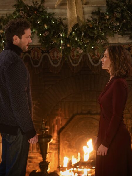 Scena rozgrywająca się przy kominku ozdobionym świątecznymi girlandami i złotą kokardą. Kobieta w bordowej sukni i mężczyzna w ciemnym swetrze stoją naprzeciw siebie w ciepłym świetle ognia, w atmosferze napięcia i niewypowiedzianych emocji.