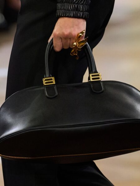 Czarna skórzana torba o zaokrąglonej formie z masywnym, złotym suwakiem i charakterystycznymi uchwytami w kształcie litery „B”. Stylowy kontrapunkt do minimalistycznego, czarnego looku, który podkreśla monumentalny charakter kolekcji Oliviera Rousteinga.