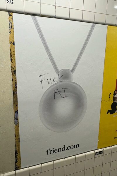 Naszyjnik AI: Reklama z kampanii Friend w metrze, przedstawiająca zdjęcie minimalistycznego, srebrzystego wisiorka z napisem friend.com, z dopisanym na plakacie hasłem „Fuck AI” – akt sprzeciwu wobec sztucznej inteligencji i komercyjnych technologii.