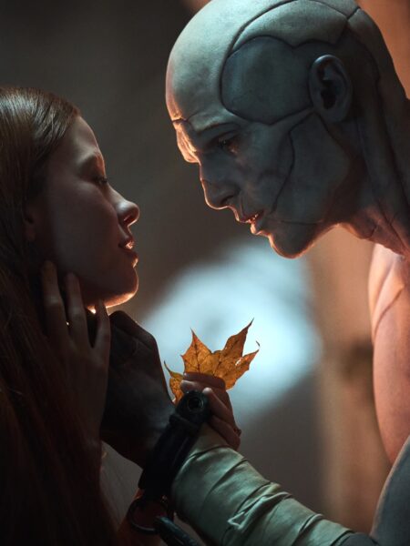 Intymne ujęcie ukazujące zbliżenie między kobietą a bladym, humanoidalnym stworzeniem o kamiennej skórze i bliznach. Postać trzyma w dłoni złoty liść, który staje się symbolem kruchości i czułości w kontrze do mrocznego tła.