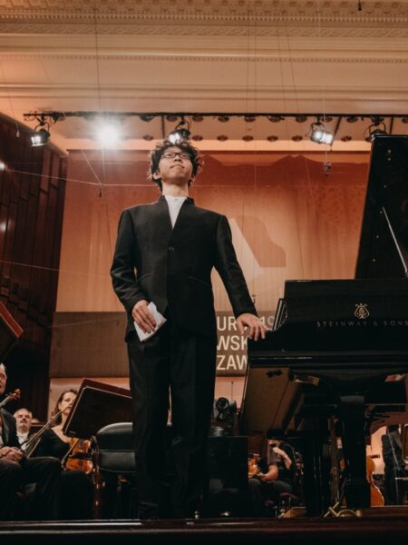 Młody pianista w czarnym garniturze stoi obok fortepianu Steinway & Sons na scenie Filharmonii Narodowej w Warszawie. Na twarzy ma skupienie i dumę, w tle widać orkiestrę i sceniczne oświetlenie konkursu Chopinowskiego.