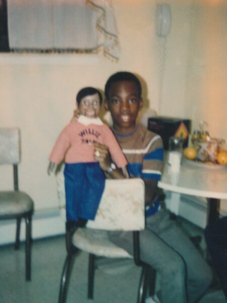 Zdjęcie w stylu retro przedstawiające chłopca trzymającego kukiełkę z szerokim uśmiechem. Kadr o ciepłej, nostalgicznej kolorystyce, wykonany we wnętrzu mieszkania z lat 80.