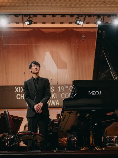 Pianista w eleganckim, czarnym garniturze stoi przy fortepianie Fazioli podczas występu w Filharmonii Narodowej. Za nim widoczny jest baner Konkursu Chopinowskiego oraz reflektory sceniczne tworzące podniosły nastrój.