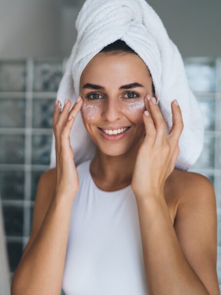 Młoda, uśmiechnięta kobieta z ręcznikiem na głowie nakłada krem ​​nawilżający na twarz, stojąc w domowej łazience. 30-letnia, szczęśliwa kobieta wykonuje codzienne poranne rytuały. Ciesz się zdrową skórą dzięki pielęgnacji kolagenem.