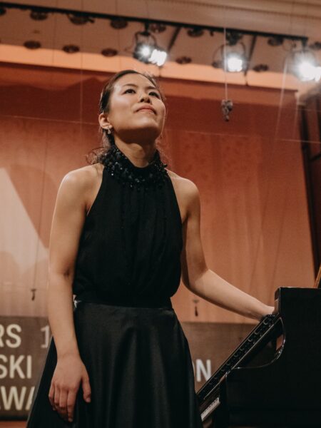 Pianistka w czarnej, eleganckiej sukni kłania się po występie na scenie Konkursu Chopinowskiego. Jej wyraz twarzy łączy wdzięk, wzruszenie i satysfakcję z występu, a w tle widać salę Filharmonii Narodowej.