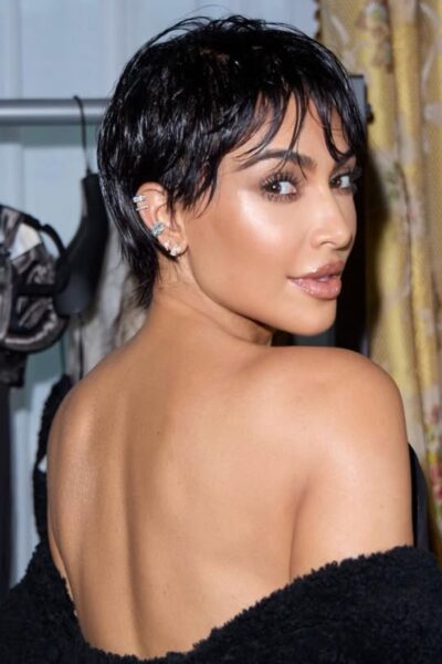 Pixie cut Kim Kardashian: Kobieta w czarnej stylizacji, odwrócona lekko plecami do obiektywu. Jej krótkie włosy w stylu wet look kontrastują z odsłoniętymi plecami, a na uszach widać srebrne kolczyki i nausznice. W tle widoczny stojak z eleganckimi ubraniami.