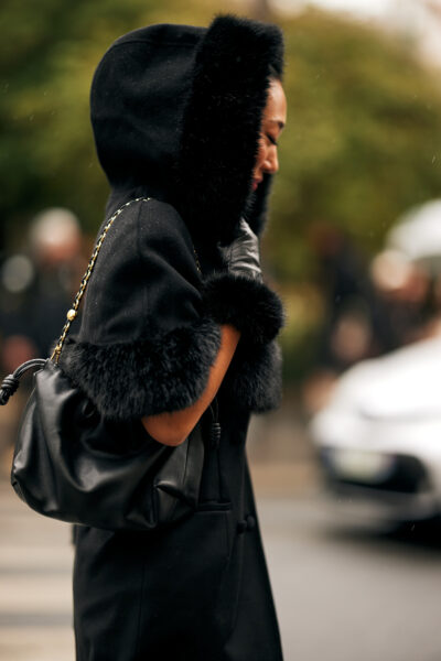 Trendy na jesień zimę 2025: Stylizacja street style z Paryża – kobieta w czarnym płaszczu z obszernym kapturem wykończonym futrzanym obszyciem, do którego dopasowane są mankiety. Całość dopełnia czarna skórzana torebka na złotym łańcuszku i czarne skórzane rękawiczki. Elegancka, zimowa stylizacja uchwycona podczas deszczowego dnia.