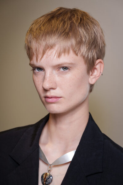 Kobieta o jasnych włosach w krótkiej, geometrycznej fryzurze. Jej twarz zdobi naturalny makijaż, a całość uzupełnia ciemna marynarka i charakterystyczny naszyjnik z metalową obręczą i kamieniem o nieregularnym wzorze.