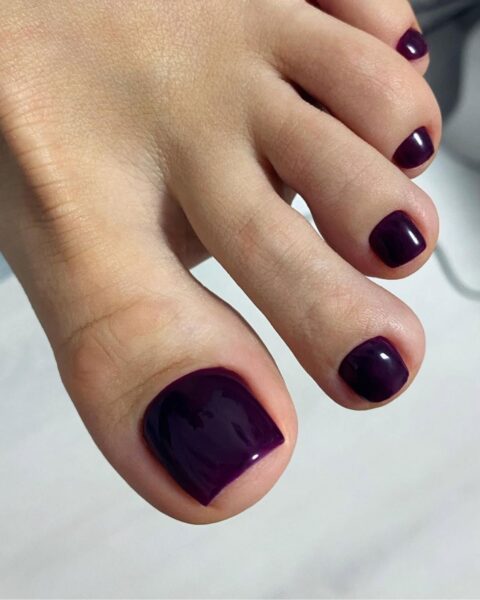 Zdjęcie przedstawia zbliżenie na zadbane stopy z perfekcyjnym pedicure'em. Paznokcie są pomalowane błyszczącym, głębokim lakierem w kolorze ciemnej śliwki lub purpury, idealnie pokrywającym płytkę. Ujęcie ukazuje staranność i precyzję wykonania stylizacji.