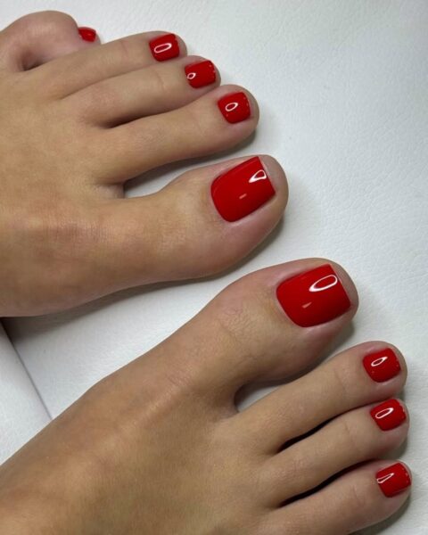 Zdjęcie przedstawia parę stóp z perfekcyjnym, klasycznym pedicure'em. Paznokcie są pomalowane na intensywny, błyszczący odcień głębokiej czerwieni. Stopy są ułożone na białej, pikowanej powierzchni.